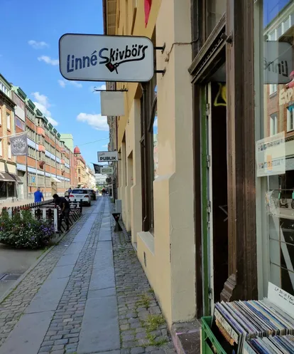 Andra Långgatan sedd längs trottoaren — Linné Skivbörs utskjutande skylt över entrén och skivbackar utanför fönstret